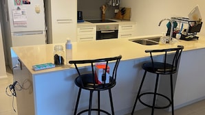 A modern kitchen with a central island, two bar stools, a refrigerator with photos, and a dish rack.