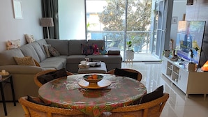 A modern living room with a sofa, a dining table with wicker chairs, a ceiling fan, and a view through large windows.