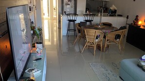 A modern living room with a flat-screen TV, a dining area with wicker chairs, and a kitchen with white cabinets.