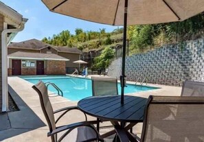 Una piscina con sillas de descanso y una sombrilla, rodeada por un muro de ladrillo y una ladera.