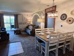 A living room with a dining table set for four, a sofa, a lamp, and a wall mural.
