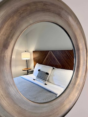 A circular mirror reflecting a bed with a wooden headboard, a bedside lamp, and a patterned pillow.