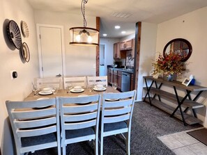 Ein Essbereich mit einem Holztisch und weißen Stühlen, einer Pendelleuchte sowie einer Küche im Hintergrund.
