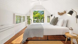 Une chambre avec un lit, un ventilateur de plafond, une fenêtre avec des stores, une petite table avec une lampe et une vue sur la verdure à l’extérieur.