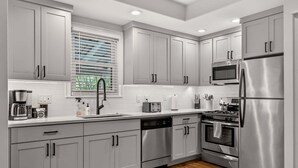 Une cuisine moderne avec des meubles gris, des appareils en acier inoxydable, un lave-vaisselle et une fenêtre avec des stores.