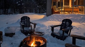 A cozy outdoor seating area with a fire pit, surrounded by snow-covered ground, and a building with large windows in the background. - Chalet Saison | Nature • Sauna - Hiking - Softness (Chertsey)