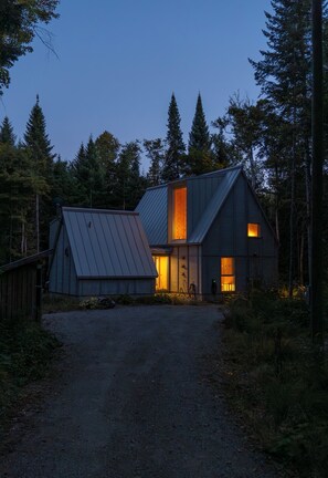 A rustic cabin with a sloped roof, a large window, and a door, surrounded by a forested area. - Chalet Saison | Nature • Sauna - Hiking - Softness (Chertsey)