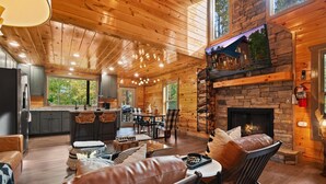 A cozy cabin interior with a stone fireplace, wooden ceiling, and a dining area with a view of the outdoors.