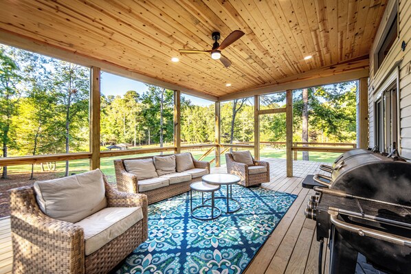 A covered outdoor area with a wooden ceiling, large windows, a barbecue grill, wicker furniture, and a patterned rug.