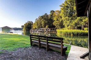 Un banco de madera junto a un lago tranquilo, con un muelle que se adentra en el agua, rodeado de vegetación exuberante y un edificio pequeño al fondo.