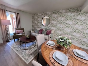 Un salon avec un canapé, une table basse, une table à manger dressée avec de l’argenterie et un papier peint à motifs floraux.