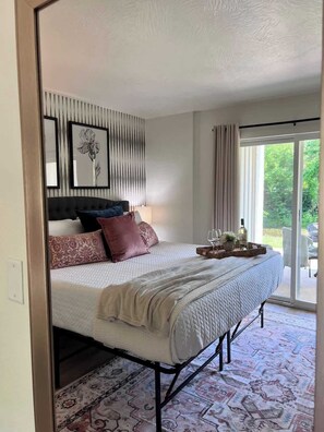 A bedroom with a bed, pillows, a bedside table with a lamp, a tray with glasses and a bottle, and a view of greenery through a window.