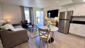 Una sala de estar moderna con un sofá, una mesa de comedor con sillas, una zona de cocina con refrigerador y un televisor montado en la pared.