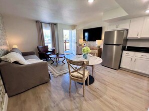 A modern living room with a sofa, a dining table with chairs, a kitchen area with a refrigerator, and a TV mounted on the wall.