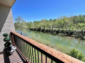 A balcony with a wooden railing overlooking a river, surrounded by greenery and trees.