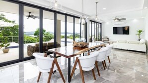A modern dining area with a large table and white chairs, large windows with a view of greenery, and ceiling fans. - Flamingo Luxury Makai Villa Next to the Beach (Playa Flamingo)