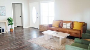 A living room with a brown leather sofa, a white coffee table, and a green armchair. There is a large window with a view of the outside, a door, and a small table with a plant. - Dothan Westside Retreat (Dothan)