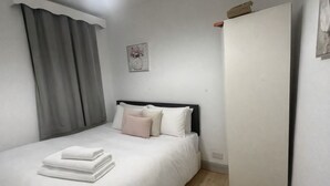 Una cama bien tendida con sábanas blancas y una toalla doblada, una cortina gris y un cuadro en la pared.