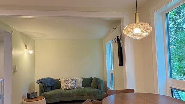 Ein modernes Wohnzimmer mit einem grünen Sofa, einem runden Holzesstisch mit Stühlen, einer Pendelleuchte und großen Fenstern mit Blick auf Bäume.