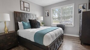 A bedroom with a large bed, a nightstand, a dresser, and a window with blinds. - Stylish Memphis Home w/ Large Yard + Cozy Interior (Memphis)