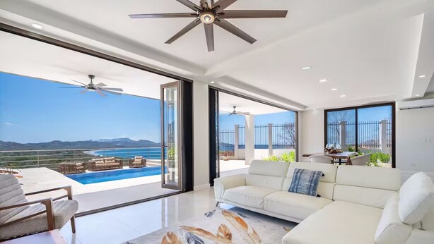 Ein geräumiger Wohnbereich mit großem Fenster, Deckenventilator und Blick auf Pool und Berge.