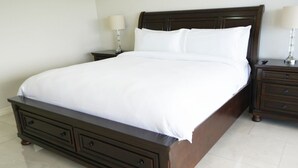 Un grand lit en bois foncé, recouvert de draps blancs, deux tables de chevet avec des lampes et un meuble à tiroirs placé sous le lit.