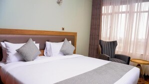 A hotel room with a large bed, a wooden headboard, two grey patterned pillows, a grey upholstered chair, and a small round table. - TJ'S Boutique Hotel Eldoret (Eldoret)