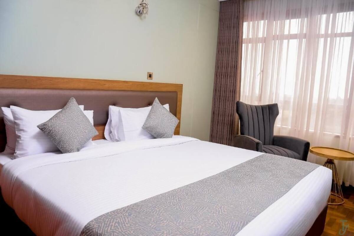 A hotel room with a large bed, a wooden headboard, two grey patterned pillows, a grey upholstered chair, and a small round table.
