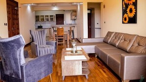 A living room with a sofa, armchairs, and a coffee table. There is a kitchen in the background with cabinets and a dining area. - TJ'S Boutique Hotel Eldoret (Eldoret)