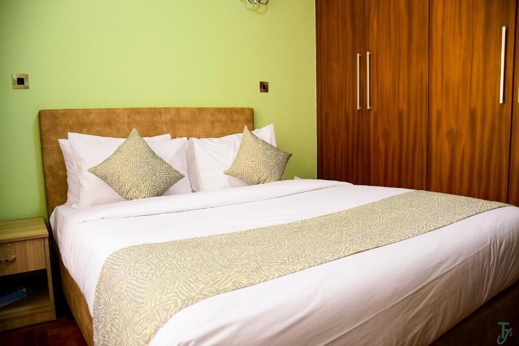 Una cama bien hecha con colcha y almohadones estampados, un armario de madera con tiradores y una mesita de noche con un cajón.