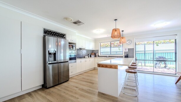 A modern kitchen with stainless steel appliances, a large island with bar stools, and a view of the backyard through a sliding glass door. - Under the Grapevines with Pool & BBQ (Dubbo)
