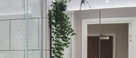 A modern bathroom with a glass shower enclosure, a marble-tiled wall, a sink with toiletries, and a hanging plant.