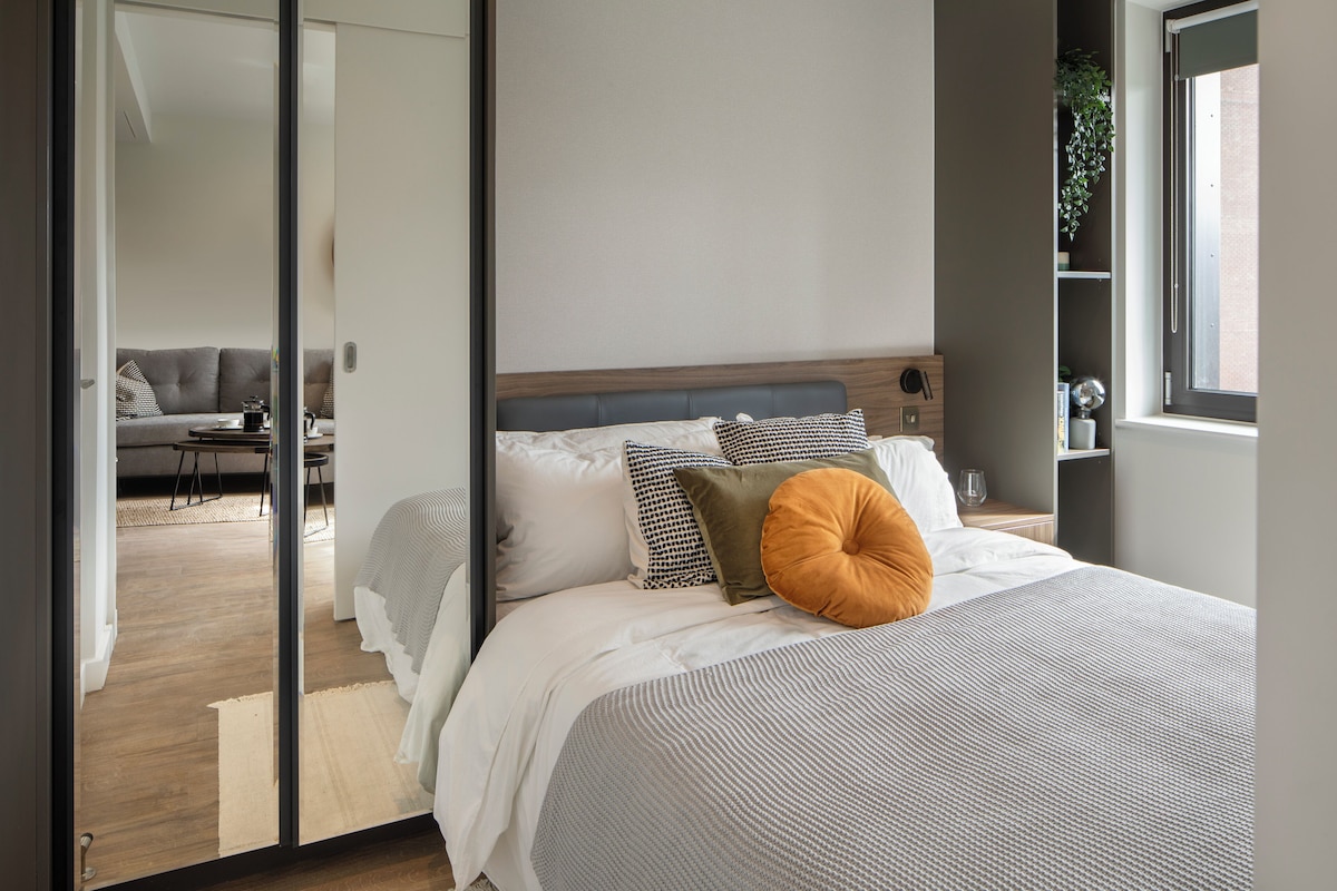 A modern bedroom with a large bed, a wooden headboard, and a view of a living area through an open door.