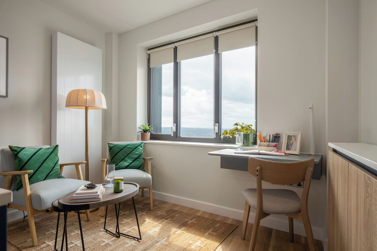 A cozy room with a desk, two chairs, a lamp, and a view of the sea through a window.