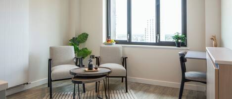 Una sala de estar moderna con dos sillones, una mesita redonda, una planta en maceta y vista a la ciudad a través de un ventanal grande.
