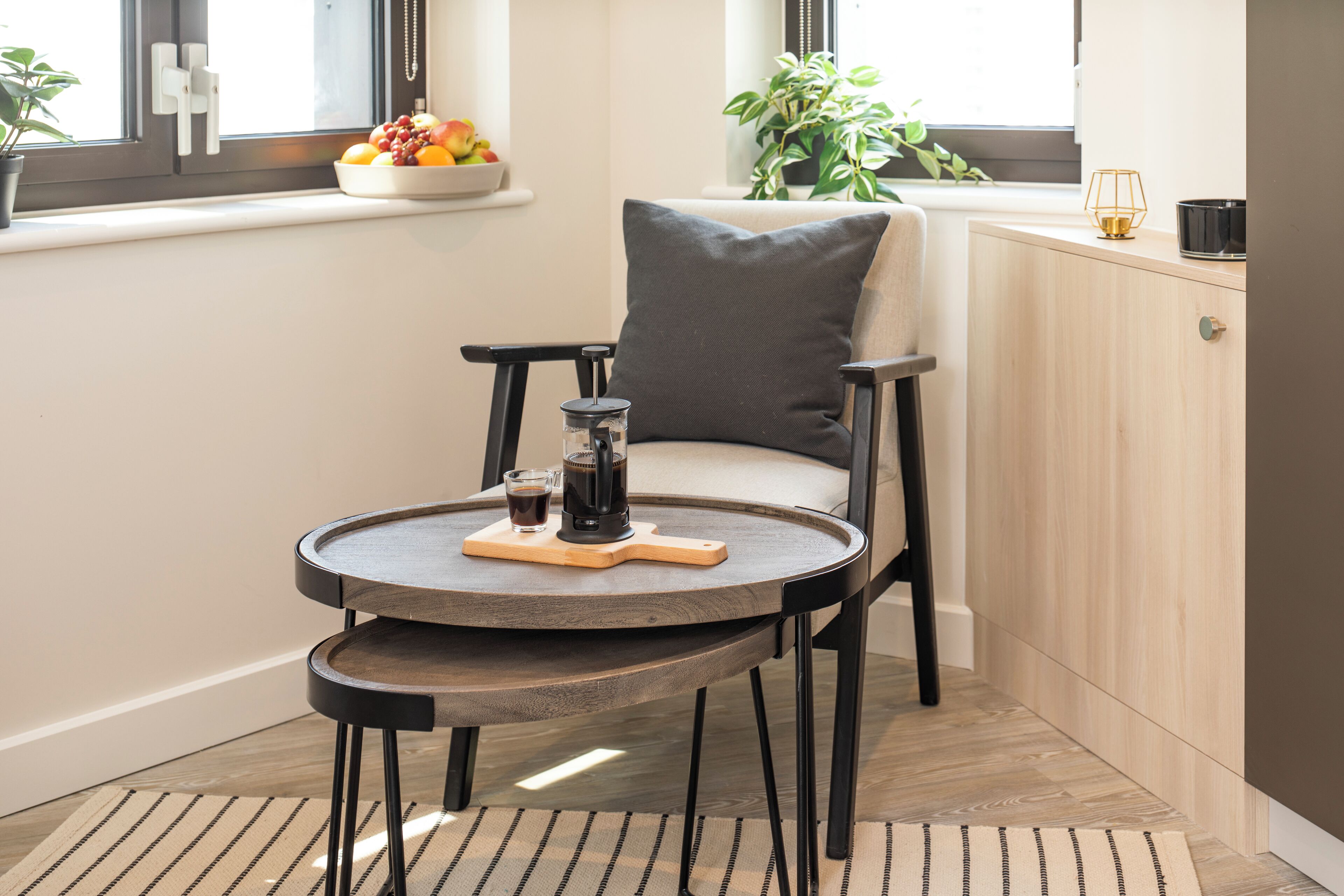 A cozy corner with a round table, a chair, a coffee maker, and a bowl of fruit on a windowsill.