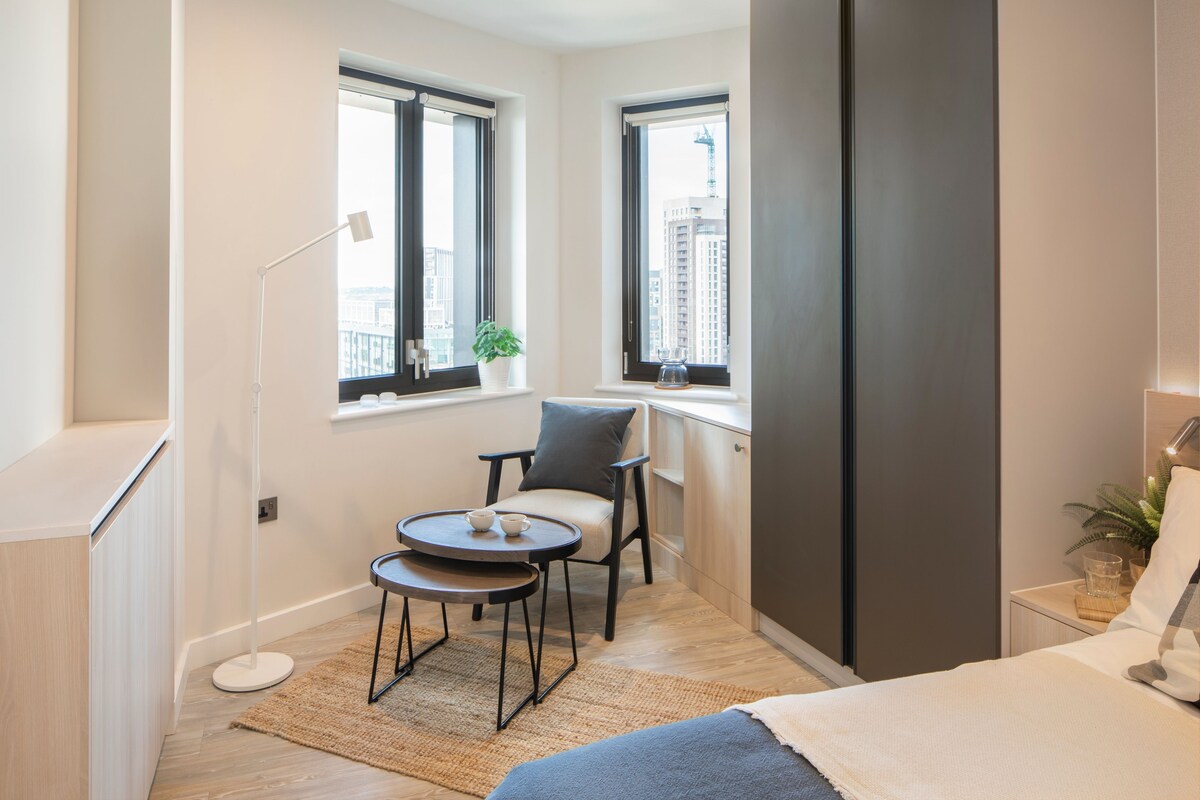 A compact room with a bed, a small table with two chairs, a floor lamp, and a window with a view of buildings.