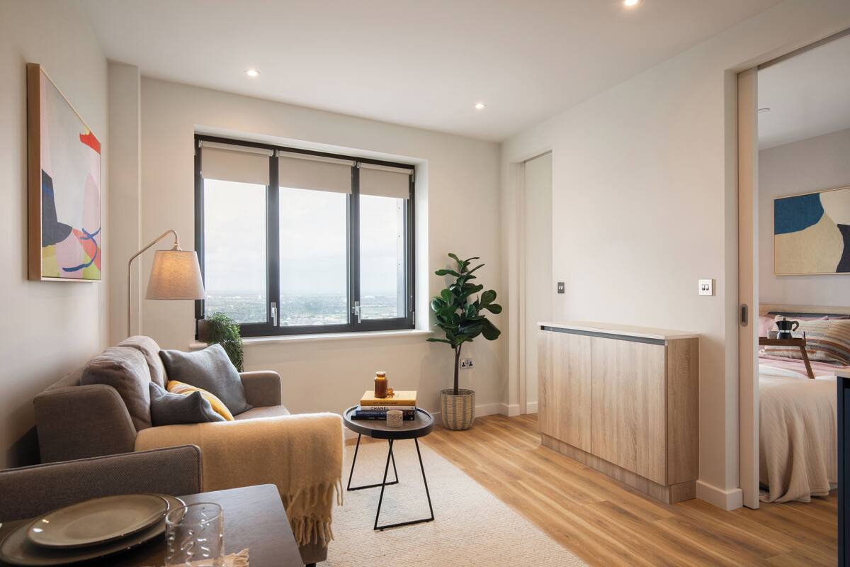 A modern living room with a sofa, a small table with a lamp, a large window, and a view of the city.