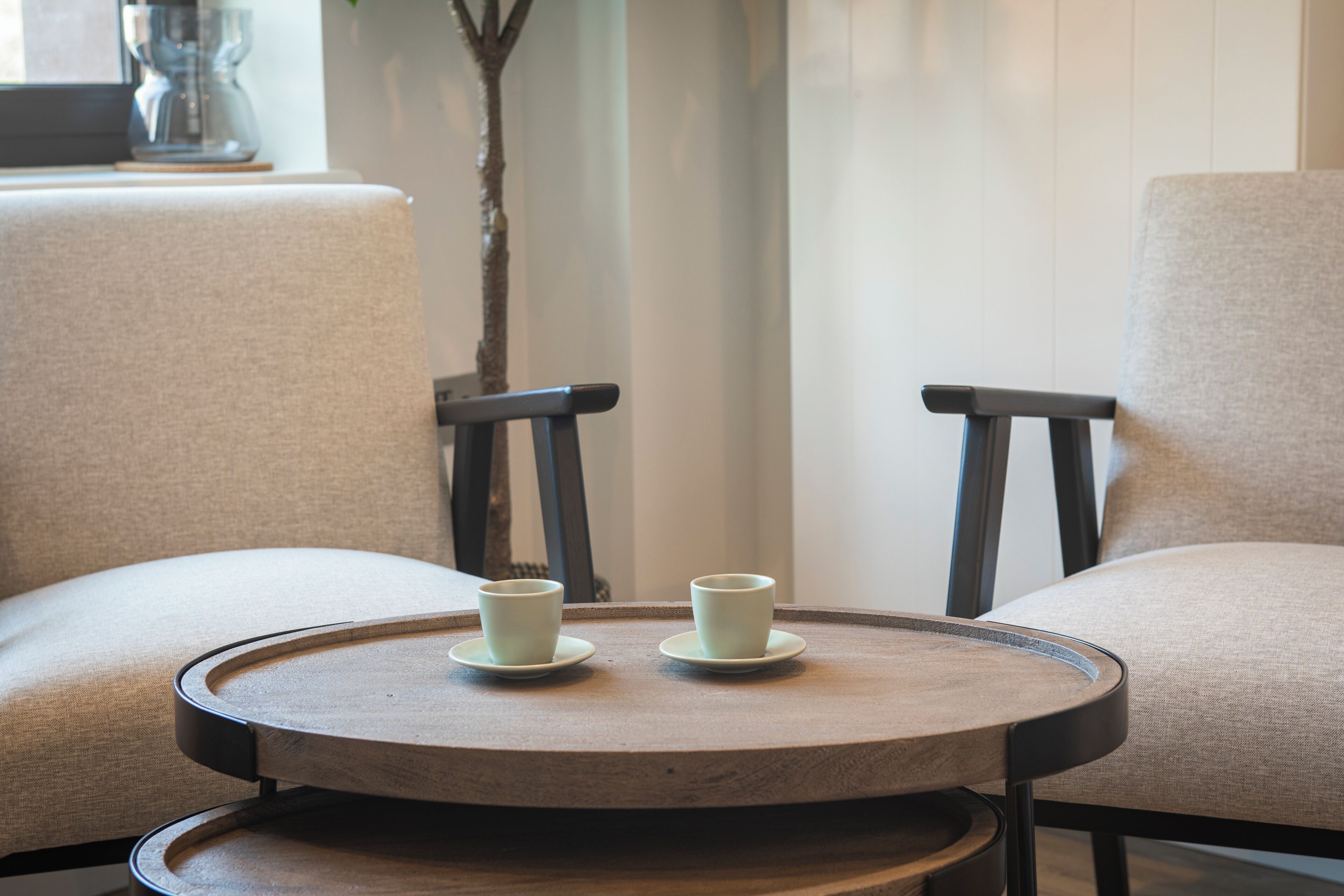 A round wooden table with two white cups on saucers, flanked by two upholstered chairs with dark wooden legs.