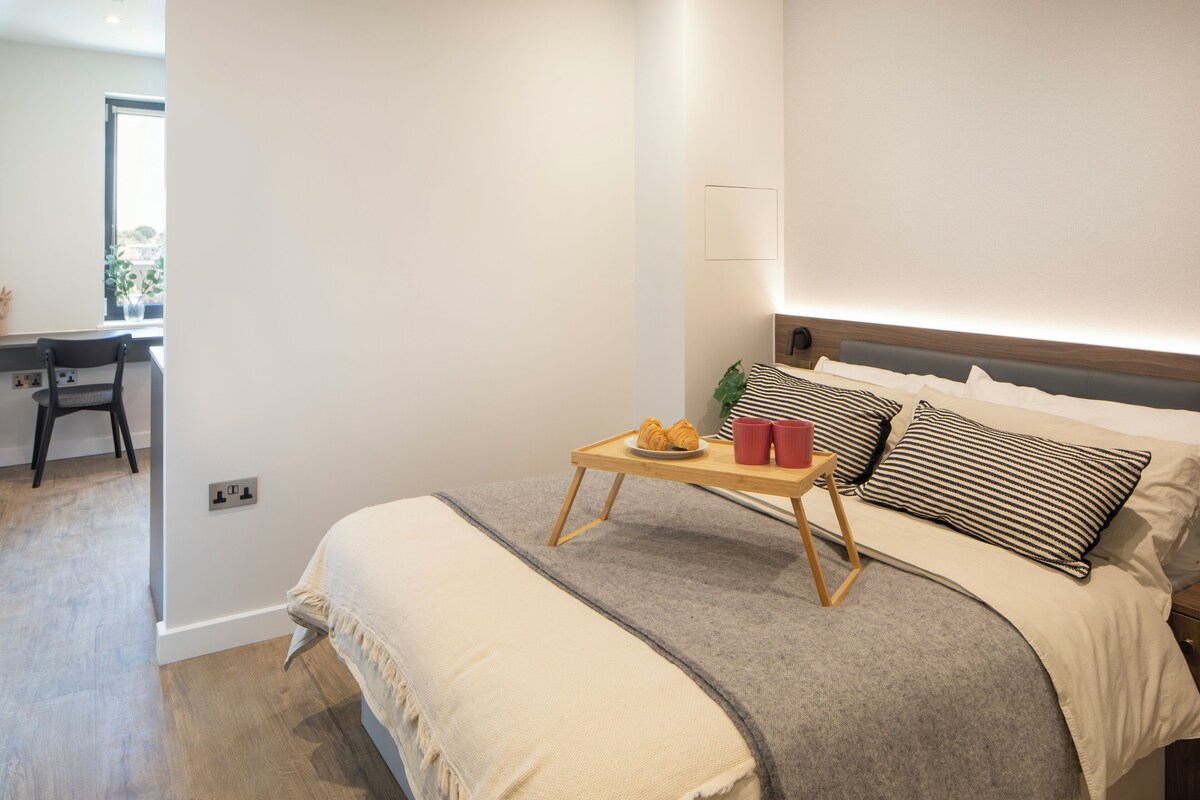 A bedroom with a bed, bedside table with a tray of croissants and cups, a desk, and a chair.