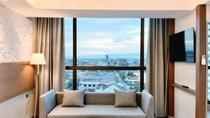 A modern hotel room with a large window, a sofa, a small table, and a flat-screen TV. - The Jogja Hotel & Conference Center - Malioboro (Yogyakarta)