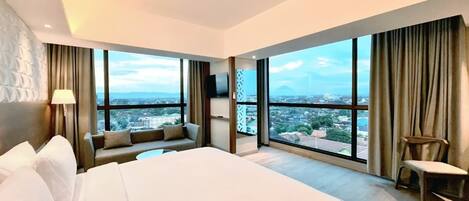 Una habitación de hotel moderna con una cama grande, un sofá, una silla y un televisor. La habitación tiene ventanas amplias que ofrecen vistas a la ciudad.