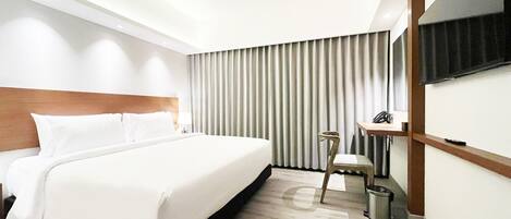A modern hotel room with a large bed, a desk, a chair, a TV mounted on the wall, and a window with curtains.