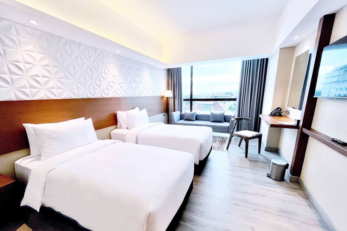 Hotelkamer met twee bedden, een slaapbank, een stoel, een bureau en een groot raam met uitzicht op de stad.