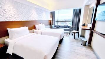 Hotelkamer met twee bedden, een slaapbank, een stoel, een bureau en een groot raam met uitzicht op de stad.