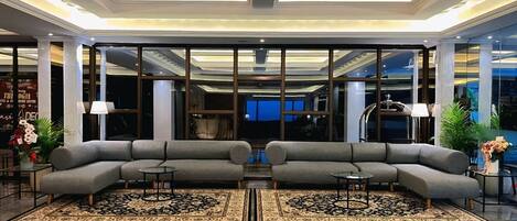A modern lobby with a grey sectional sofa, a reflective floor, and a ceiling with wooden panels and recessed lighting.