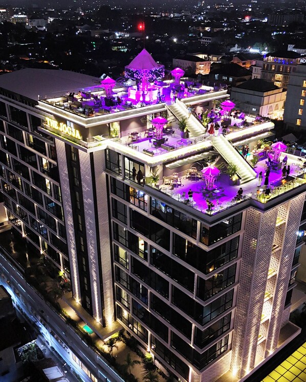 Un hotel de varios pisos con terraza en la azotea, mobiliario exterior y un escenario.
