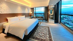 A modern hotel room with a large bed, a sofa, and a view of a cityscape through large windows. - The Jogja Hotel & Conference Center - Malioboro (Yogyakarta)