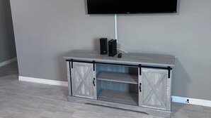 Ein Zimmer mit einem wandmontierten Flachbildfernseher, einem grauen Schrank mit kreuzförmigen Türen und einem hellgrauen Fliesenboden.