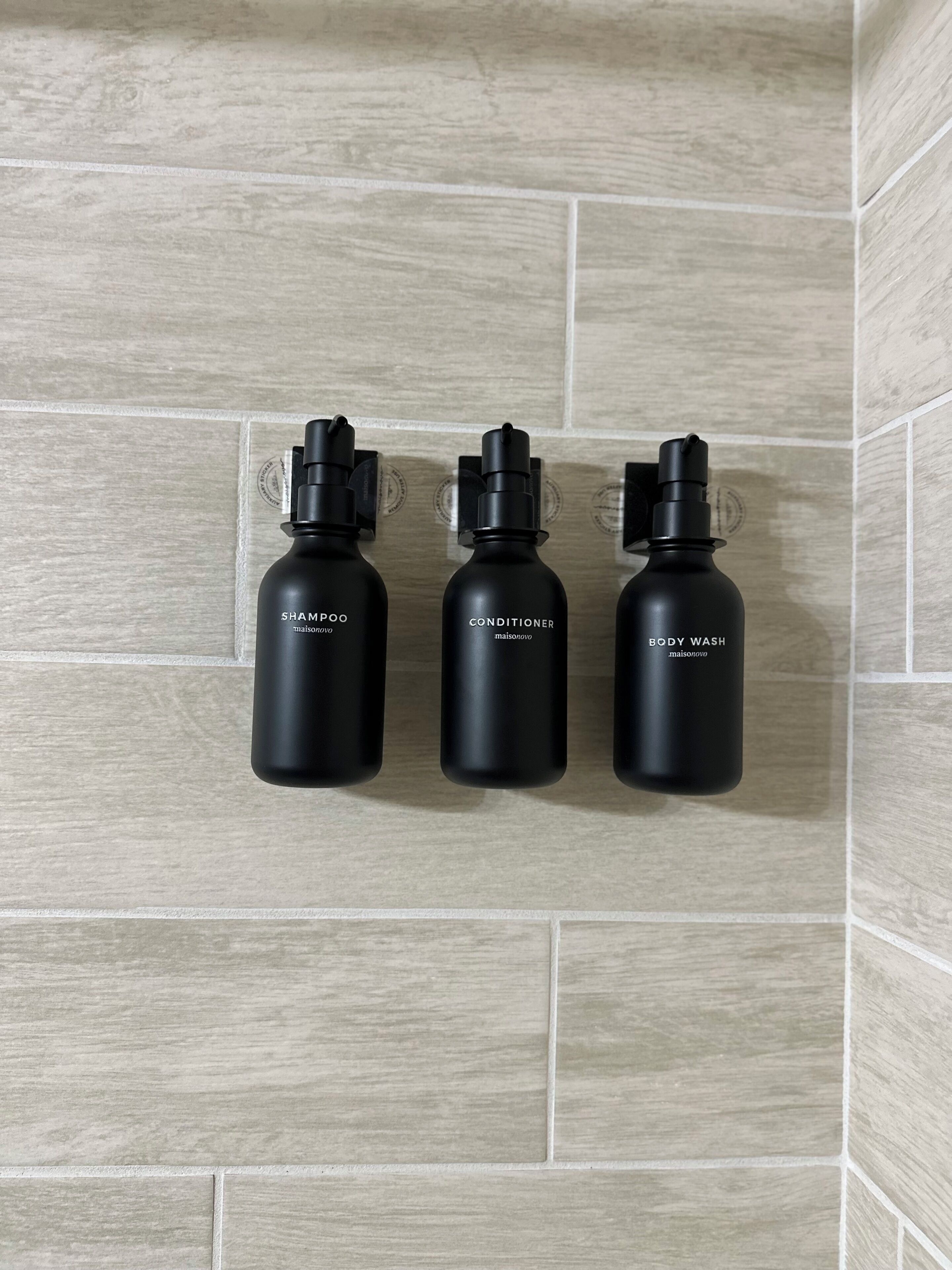 Tres botellas negras de productos maisonnovo sobre una pared de baldosas.